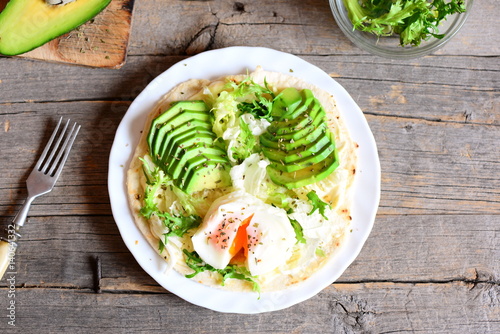 Vegetarian breakfast with a poached egg, avocado slices, chinese cabbage, lettuce, tortilla, sauce and spices. Easy avocado and poached egg tortilla on a plate and on old wooden background. Top view