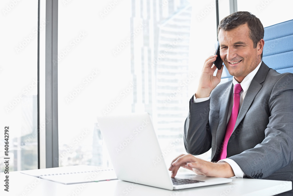Smiling mature businessman talking on cell phone while using laptop in office Smiling mature businessman talking on cell phone while using laptop in office