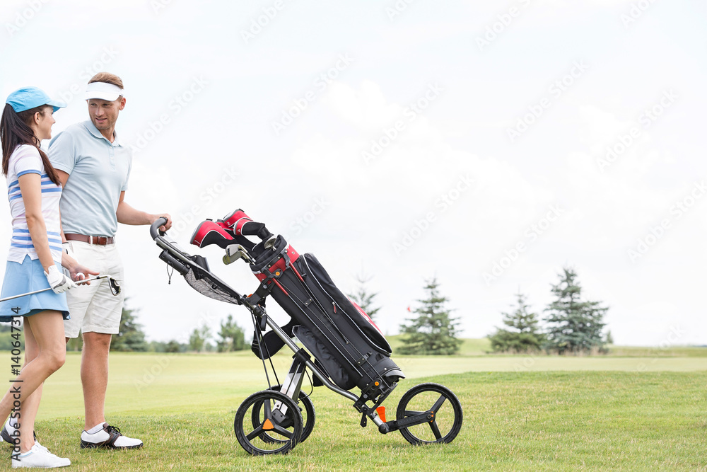 Friends with equipment talking while walking at golf course against clear sky Friends with equipment talking while walking at golf course against clear sky