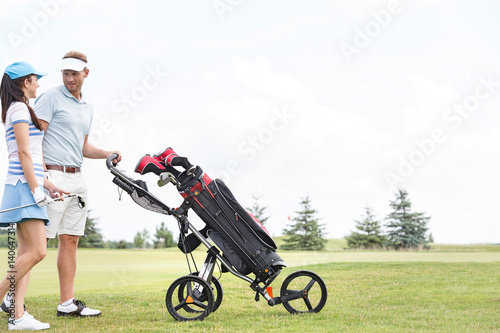 Friends with equipment talking while walking at golf course against clear sky