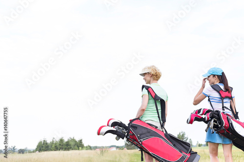 Rear view of women with golf club bags at course against clear sky