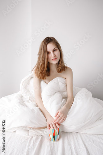 Portrait of smiling beautiful young Caucasian girl woman with long red hair sitting in bed wrapped covered with blanket, holiding cup, drinking coffee or tea, on white background.