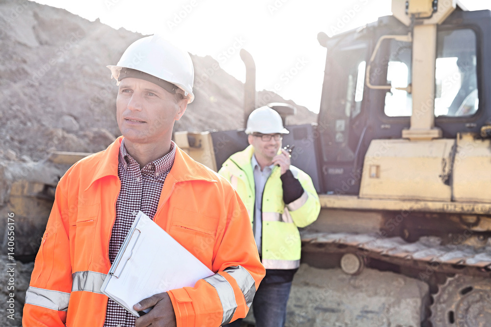 Engineer holding clipboard on construction site with colleague in background Engineer holding clipboard on construction site with colleague in background