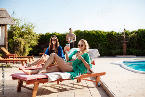 Girls smiling, drinking cocktails, sunbathing, lying near swimming pool. © Cookie Studio