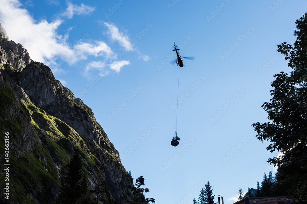 a silhouette of a helicopter carrying shipment for a mountain house in the Alps - obrazy, fototapety, plakaty 