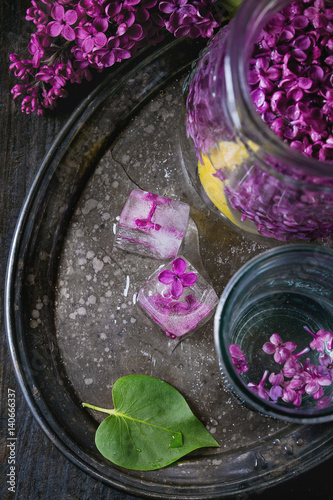 Glass and pitcher of lilac lemonade water with lemon, ice cubes with lilac flowers and lilac branch on vintage iron tray over black wooden table. Dark rustic style. Top view