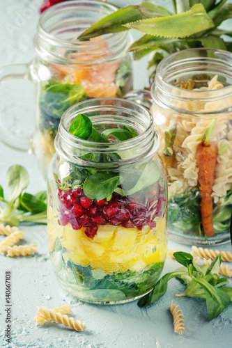 Variety of salads in mason jars. Fruit salad mango, pomegranate, greens, vegetables, wholegrain pasta, carrots and cauliflower, salmon. Standing over blue texture background. Food to go.