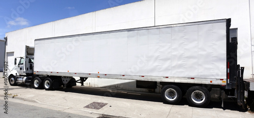 White semi freight truck with trailer parked next to white warehouse under blue sky.