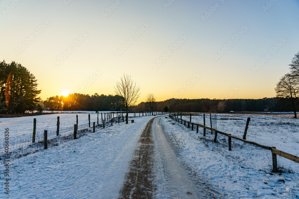 Sunset over the snow covered road through fields and forest