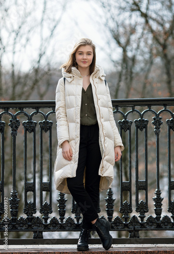 Full length posing beautiful young blonde woman standing on the spring city park in warm clothes. Cold season lifestyle freshness concept