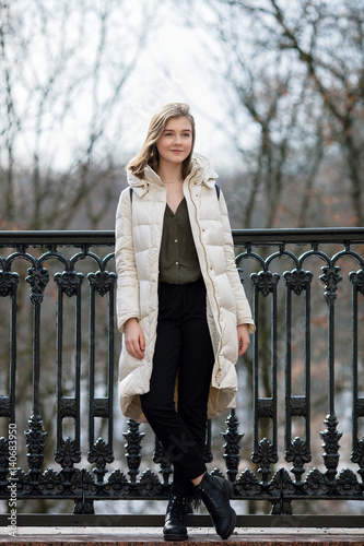 Full length posing beautiful young blonde woman standing on the spring city park in warm clothes. Cold season lifestyle freshness concept