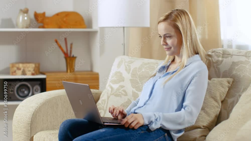 Beautiful Young Woman Sitting on a Couch in Her Living Room. She Holds Her Laptop on Her Laps and Works on It. Shot on RED EPIC-W 8K Helium Cinema Camera. Beautiful Young Woman Sitting on a Couch in Her Living Room. She Holds Her Laptop on Her Laps and Works on It. Shot on RED EPIC-W 8K Helium Cinema Camera.