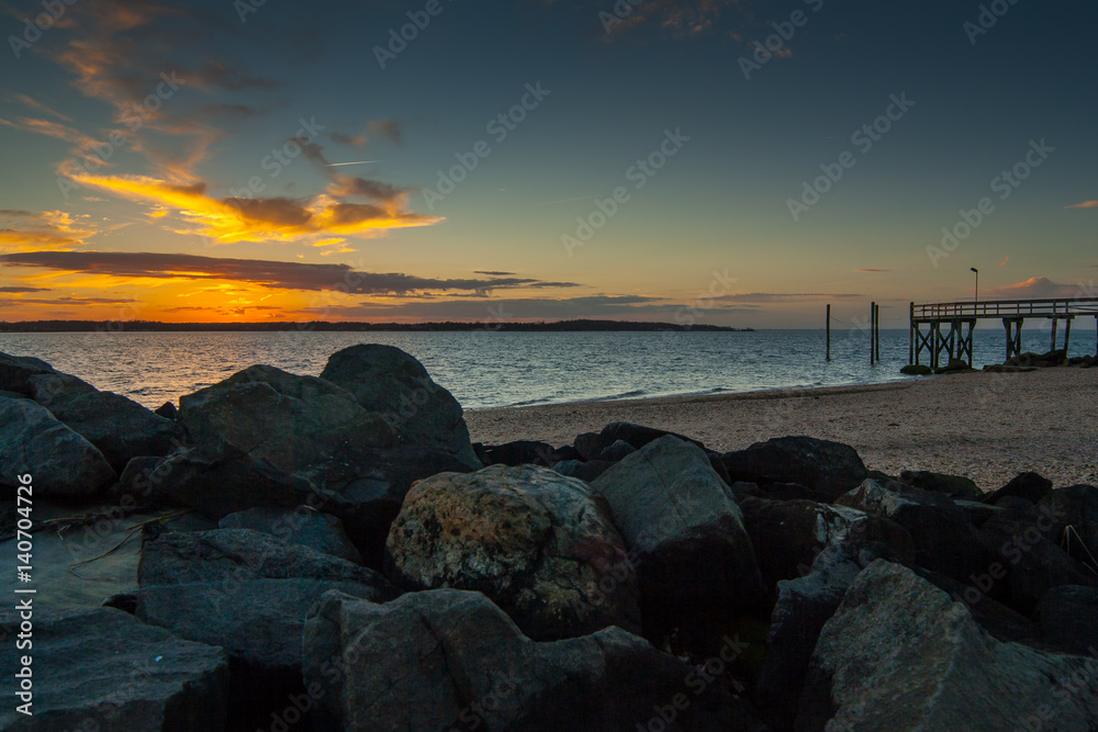 Fototapeta premium Large stones in the foreground with a rocky beach, a wooden dock, and a colorful sunset sky with clouds in the background. West Neck Beach in Huntington, NY, USA.
