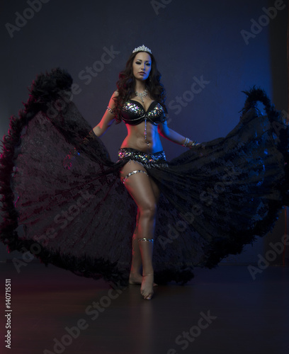 Sexy slim woman, belly dancer in black costume with crystals. Dark background with pink and blue lightening