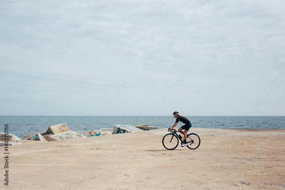 Fototapeta premium Male professional cyclist rides around a pier wearing all black near blue sea and rocky port nice light.