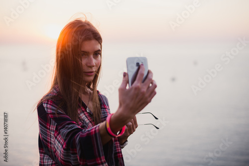 Portrait of young happy woman in hipster shirt standing in front of picturesque sea view, taking selfie with sunset or sunrise on smartphone