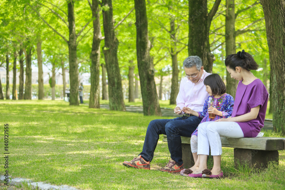 Girl sitting between mother and grandfather on park bench hands lid of drink bottle to her grandfather Girl sitting between mother and grandfather on park bench hands lid of drink bottle to her grandfather