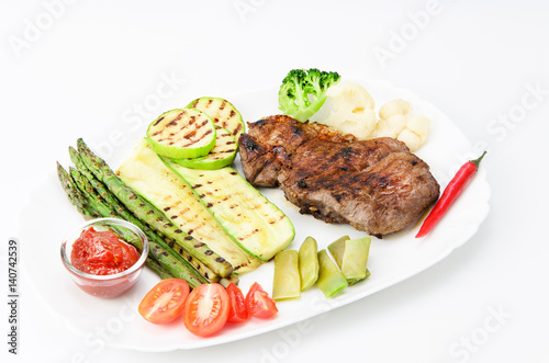 Grilled beef grilled with asparagus, zucchini, lobi, broccoli, tomato, chili and red sauce on a white plate, over white background