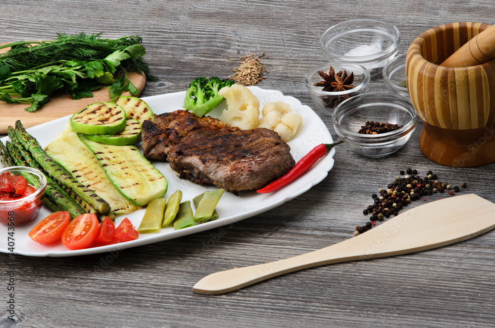 Grilled beef grilled with asparagus, zucchini, lobi, broccoli, tomato, chili and red sauce on a white plate, and other vegetables in wood background Grilled beef grilled with asparagus, zucchini, lobi, broccoli, tomato, chili and red sauce on a white plate, and other vegetables in wood background