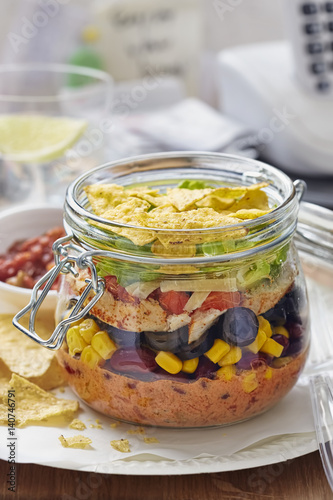 Layered Mexican sandwich jar with corn, black beans, lettuce, cheese, red peppers, lettuce and tortilla chips