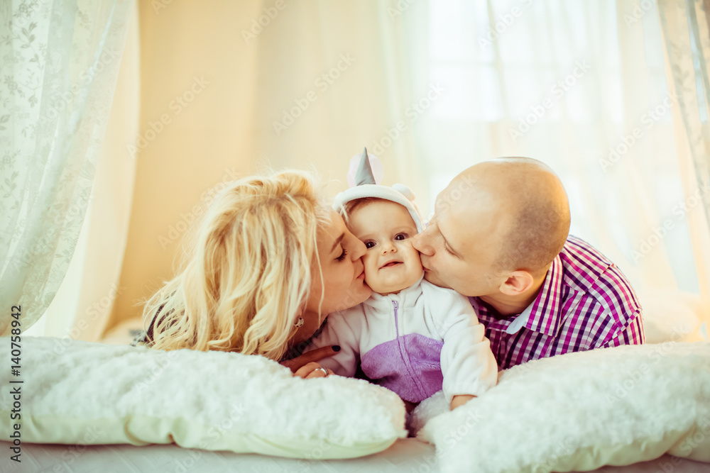 Joyful parents kiss their little daughter lying on soft bed