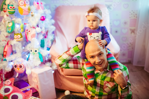 Man in bright plaid shirt holds little girl on his neck sitting before Christmas tree