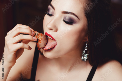 A beautiful European girl with styling and make-up bites meat. Dark background.