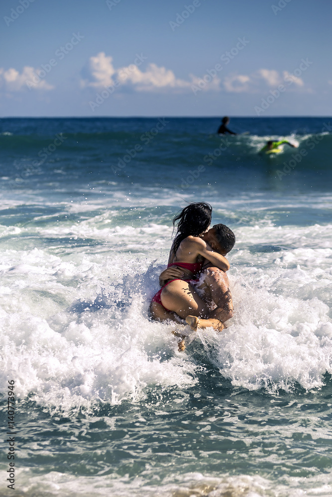 Young and attractive couple is having fun in the sea. Man is holding woman on his hands. They are waiting for wave to be covered with it. Sunny day, Barcelona, Spain Young and attractive couple is having fun in the sea. Man is holding woman on his hands. They are waiting for wave to be covered with it. Sunny day, Barcelona, Spain