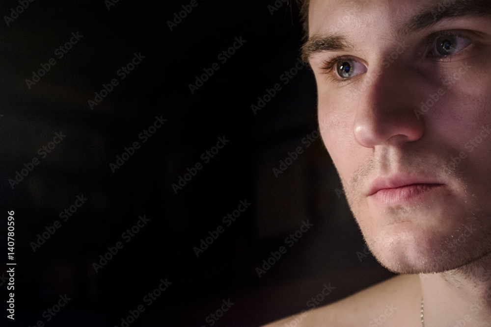 Portrait of a handsome guy with a naked torso. Photo of a close-up in a dark room and light from a lamp. Soft light. Green-eyed brunette full-face. Part of the body is the face. Place for the text.