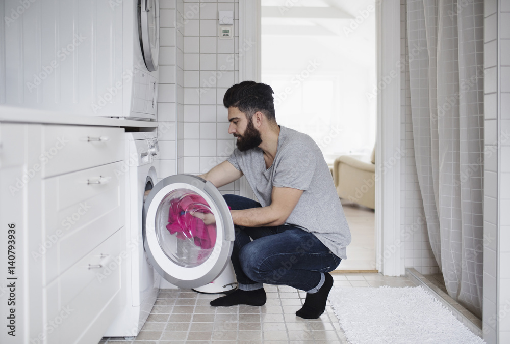 Side view of man crouching while loading clothes in washing machine at home Side view of man crouching while loading clothes in washing machine at home