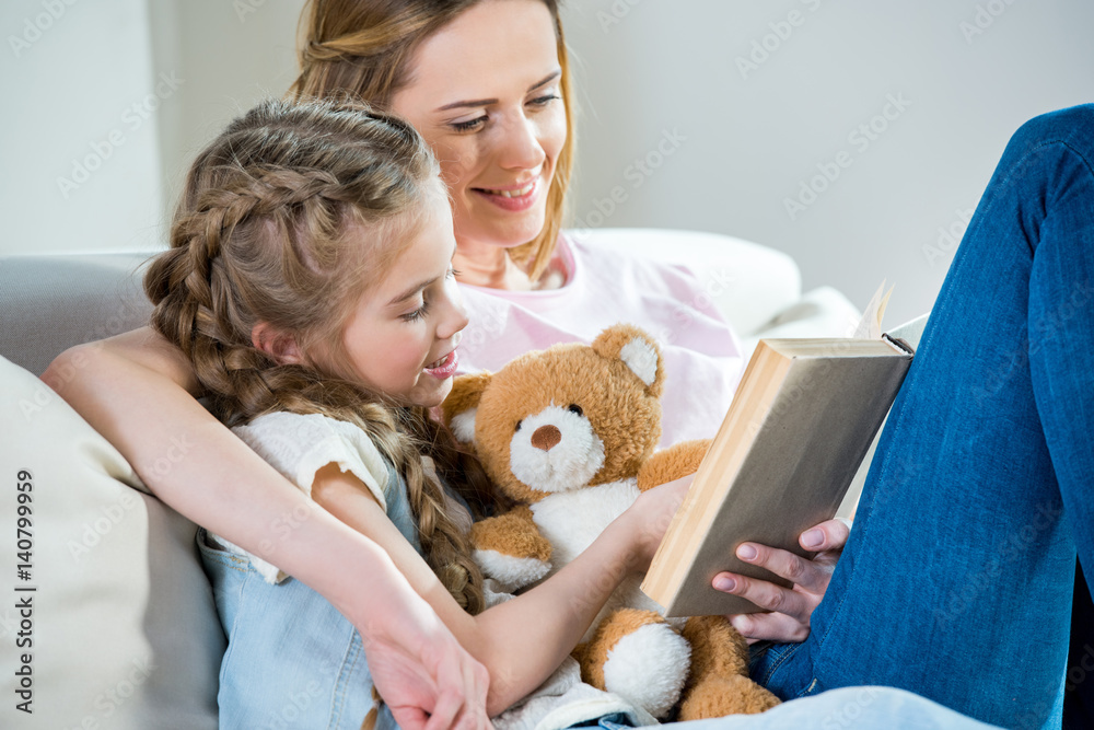 Smiling mother and daughter with teddy bear reading book on sofa Smiling mother and daughter with teddy bear reading book on sofa