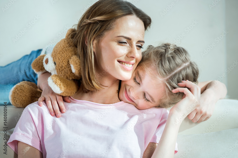 Happy mother and daughter with teddy bear hugging on sofa at home