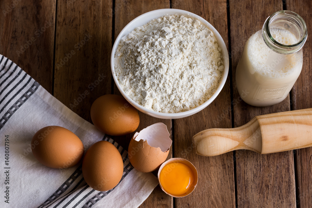 Eggs, cracked shell open yolk, flour, milk ingredients for baking, rolling pin on wood kitchen taple, towel, top view Eggs, cracked shell open yolk, flour, milk ingredients for baking, rolling pin on wood kitchen taple, towel, top view