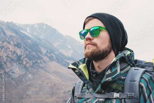 A guy with a beard and wearing sunglasses in a membrane jacket, hat, with a backpack and sticks for Nordic walking, a traveler standing and looking at the mountains
