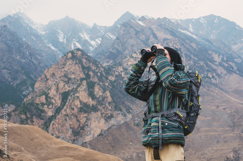 A hipster man with a beard in a hat, jacket, and a backpack in the mountains holds binoculars in his hands and looks into the distance, adventure, tourism, tracking