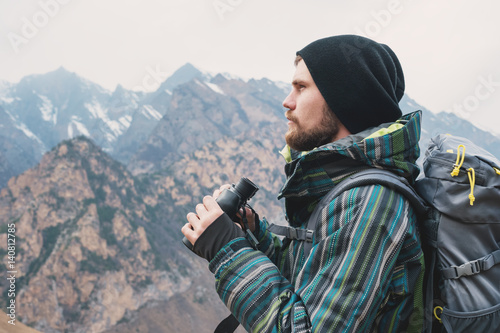 A hipster man with a beard in a hat, a jacket, and a backpack in the mountains holds binoculars, adventure, tourism, tracking