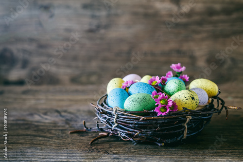 Easter background. Bright colorful eggs in nest with spring flowers over wooden dark background. Selective focus with copy space.
