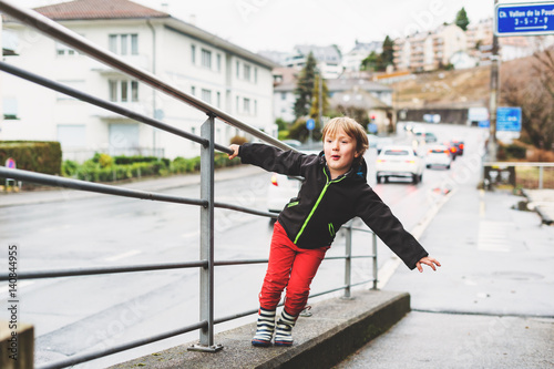 Little boy playing outdoors in early spring, walking next to road, wearing black waterproof jacket, red trousers and rain boots