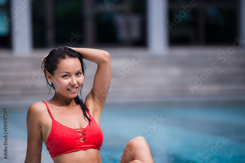 Portrait of beautiful, young lady in red bikini sitting poolside in luxury hotel