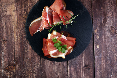 Bread with slices jamon serrano for lunch table. Sharing antipasti on party or summer picnic time over wooden rustic background.