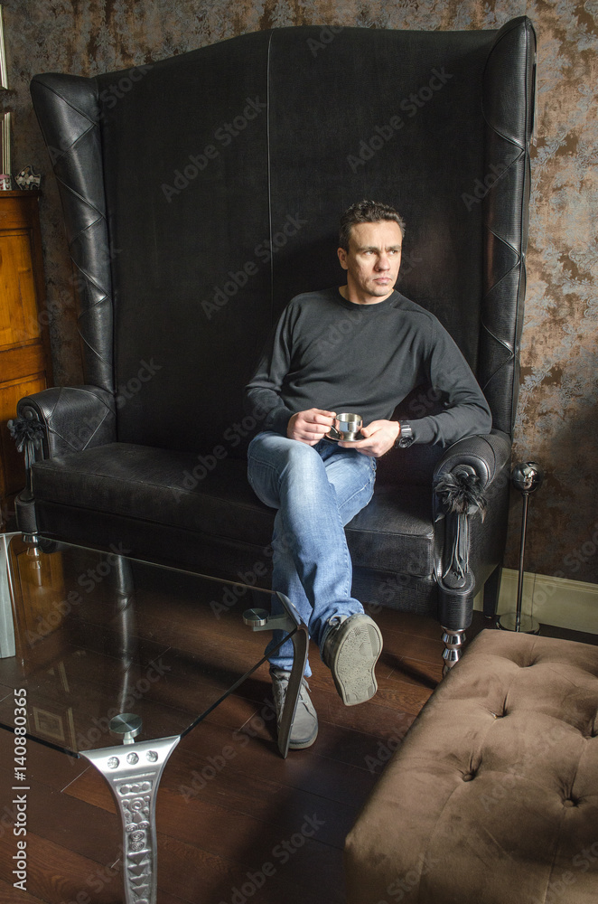 Sitting man in a black sweater and blue jeans on a black leather sofa Sitting man in a black sweater and blue jeans on a black leather sofa