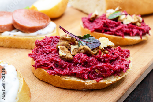 Breakfast sandwiches with boiled grated beets and walnuts on a piece of white baguette on a cutting board.