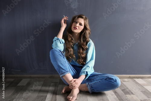 Young woman sitting on floor and smoking against color wall
