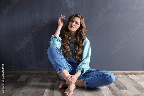 Young woman sitting on floor and smoking against color wall © Africa Studio