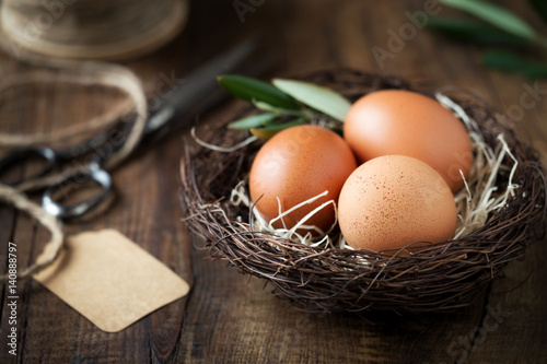 Happy Easter concept - Easter eggs in burd nest with an olive branch against dark rustic wooden background with empty gift tag sign for Happy Easter greeting message