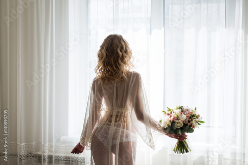 Bride near the window in her underwear. Blond sexy female Boudoir photo back to camera in front of large window. Bridal bouquet in the hand of bride. photo