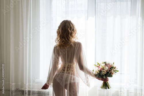 Bride near the window in her underwear. Blond sexy female Boudoir photo back to camera in front of large window. Bridal bouquet in the hand of bride.