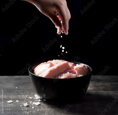 Hand sprinkling salt on raw meat of pork over black background.
