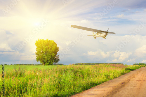 White plane flying over green field and road with a tree on the background of the sun