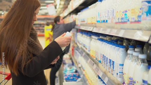 A young attractive woman is shopping in a supermarket. She is taking a bottle of milk from the shelf and puting it in a cart. 4K
